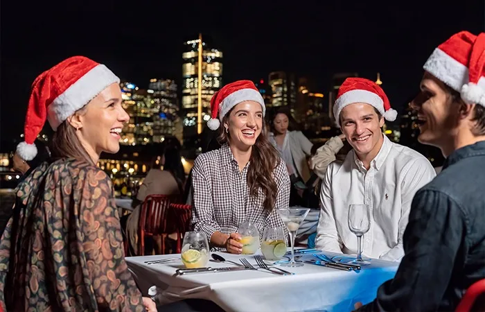 People celebrating Christmas aboard a Sydney dinner cruise, wearing Santa hats with the city skyline illuminated in the background.