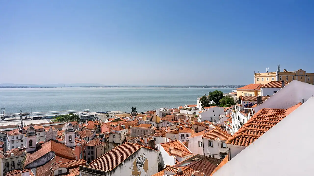 Terracotta rooftops of Lisbon's Alfama district sloping down to the wide Tagus river under a clear blue sky