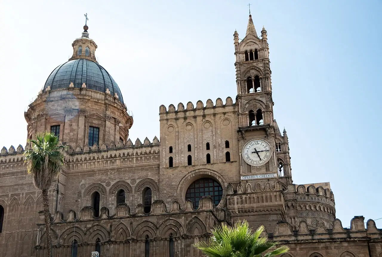 Palermo Cathedral in Sicily, with its dome, crenellated walls and Gothic clock tower, framed by palm trees under a clear sky