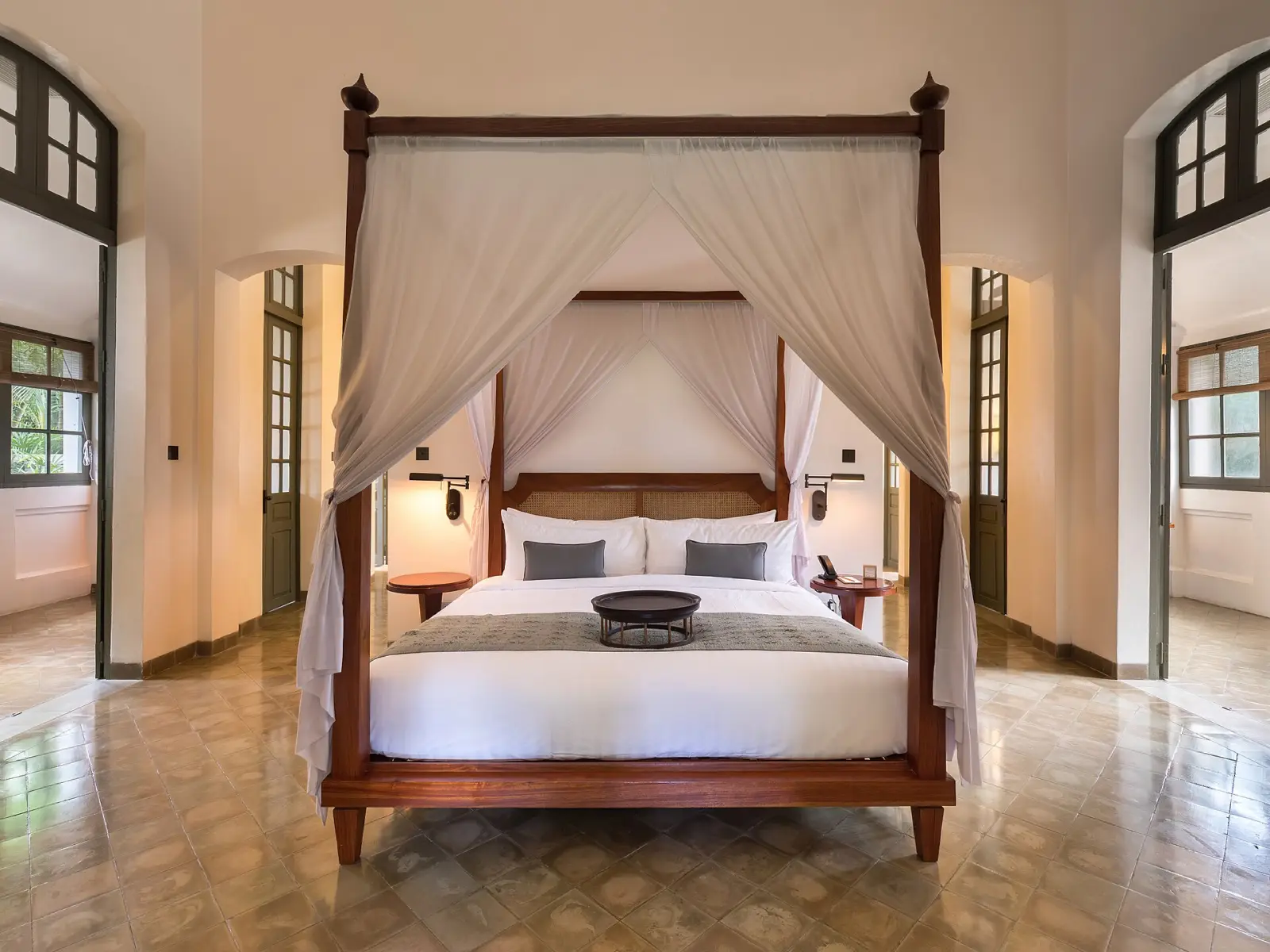 A white four-poster canopy bed with sheer curtains in a calm, symmetrical luxury hotel suite with tiled floors