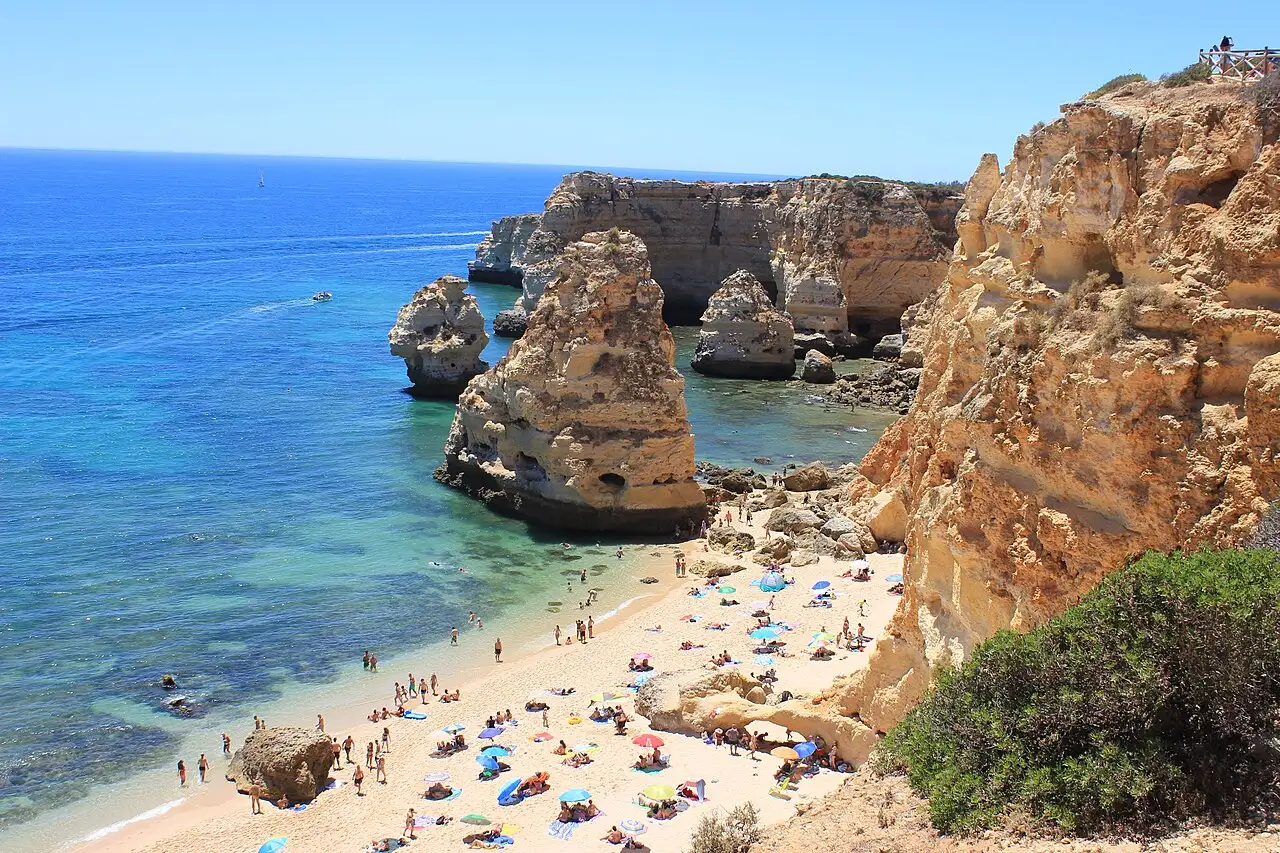 Golden limestone cliffs and sea stacks above a sandy cove with bathers and blue water at Praia da Marinha in the Algarve, Portugal