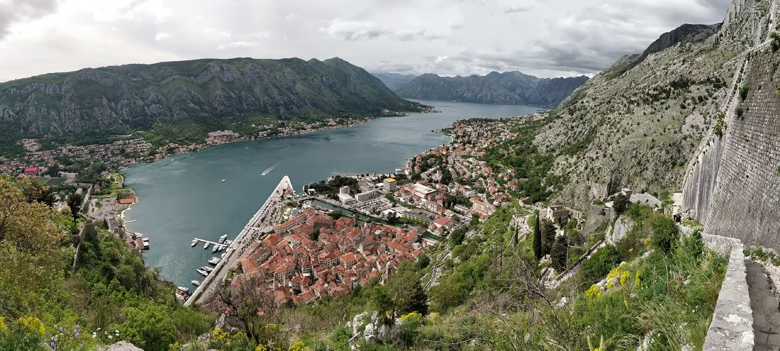 Panorama from above of Kotor’s terracotta-roofed Old Town beside the Bay of Kotor in Montenegro, with steep mountains rising on both sides of the bay